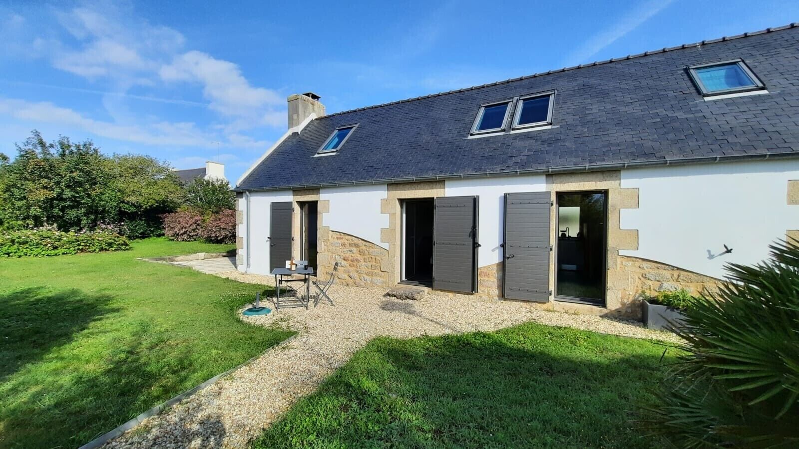A house with a sloped roof, two open doors, a small patio with a table and chairs, and a garden.