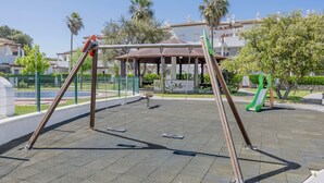 A playground with swings and a slide, surrounded by a fenced area with palm trees and buildings in the background.