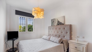 A bedroom with a bed, a television, a bedside table, a window, and a wall-mounted light fixture.