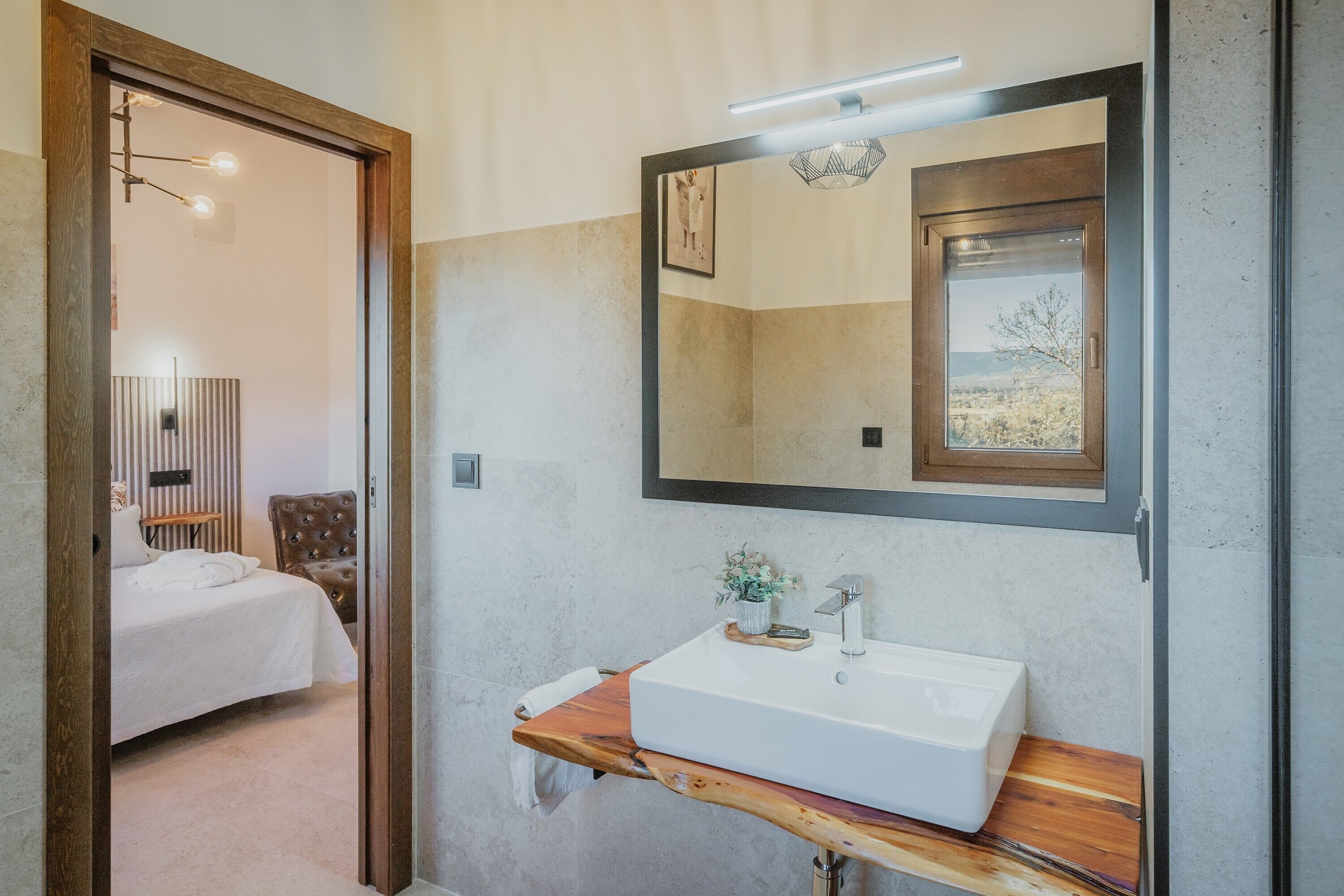 Een badkamer met een houten wastafelmeubel, een grote spiegel en uitzicht op een slaapkamer.