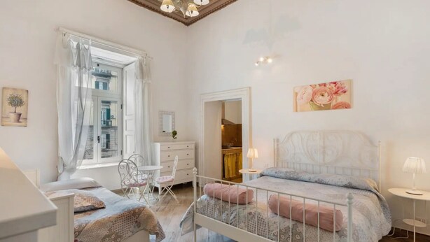 Ein Schlafzimmer mit einem Bett, einem Sessel, einem Schminktisch, einem Spiegel und einem Fenster mit Vorhängen.