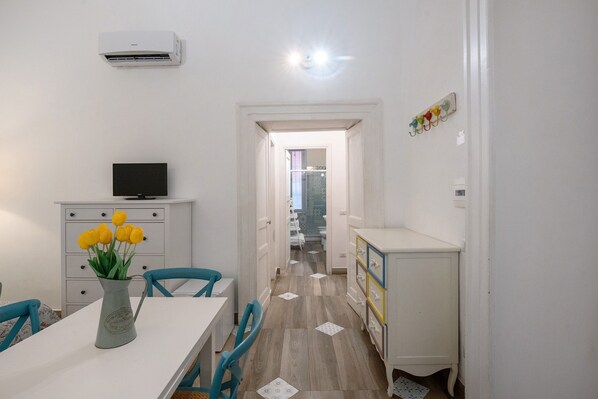 Une pièce avec un climatiseur blanc, un téléviseur à écran plat posé sur un meuble blanc, une table à manger avec des chaises et un vase rempli de fleurs jaunes.