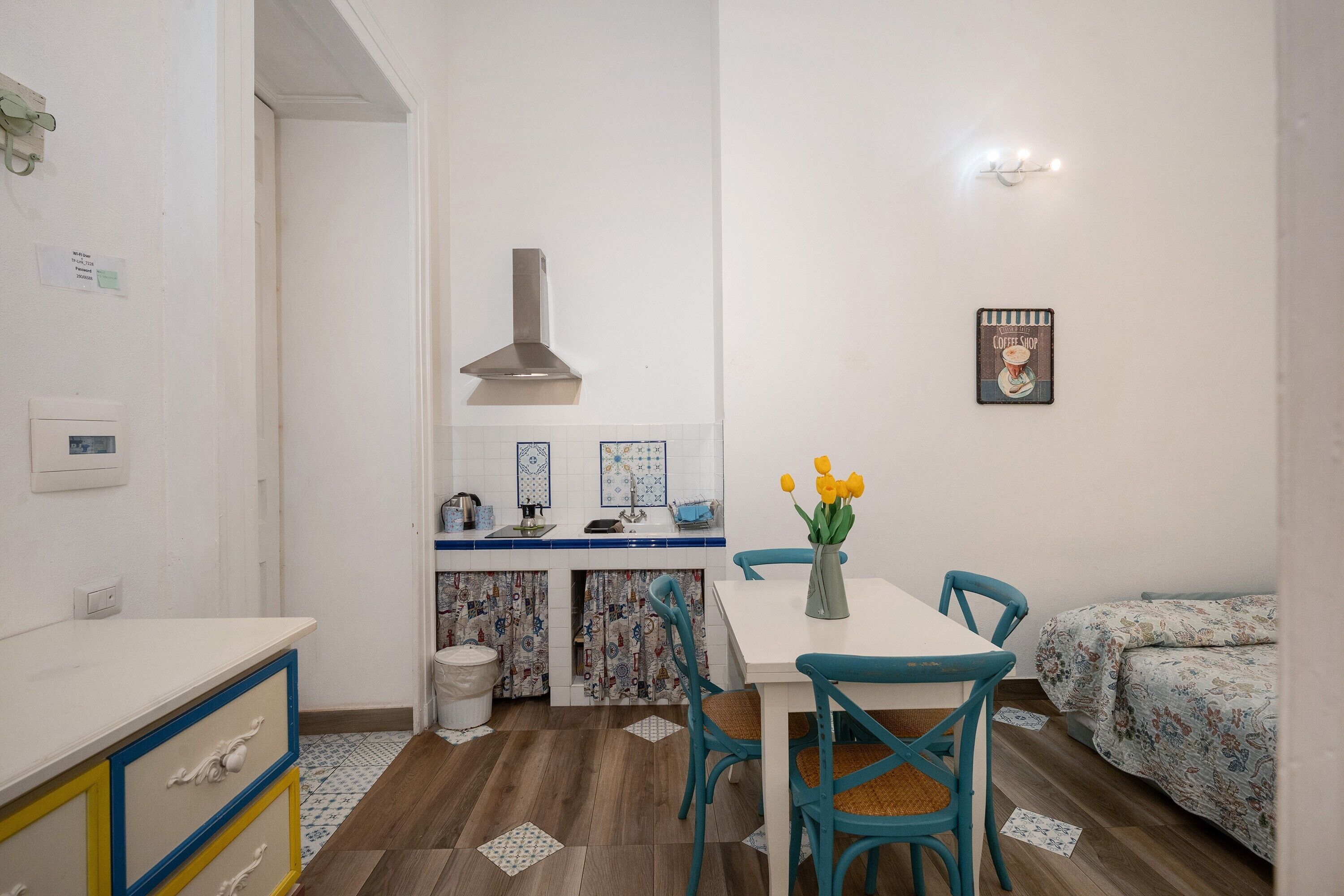 A compact kitchen with a white countertop, a sink, and a stove. There is a dining table with chairs and a vase with flowers. A bed with a floral bedspread is visible in the corner.