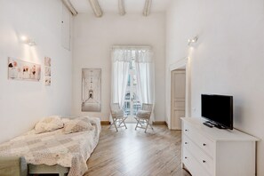 Un dormitorio con cama, dos sillas, un televisor sobre un aparador y una ventana con cortinas.