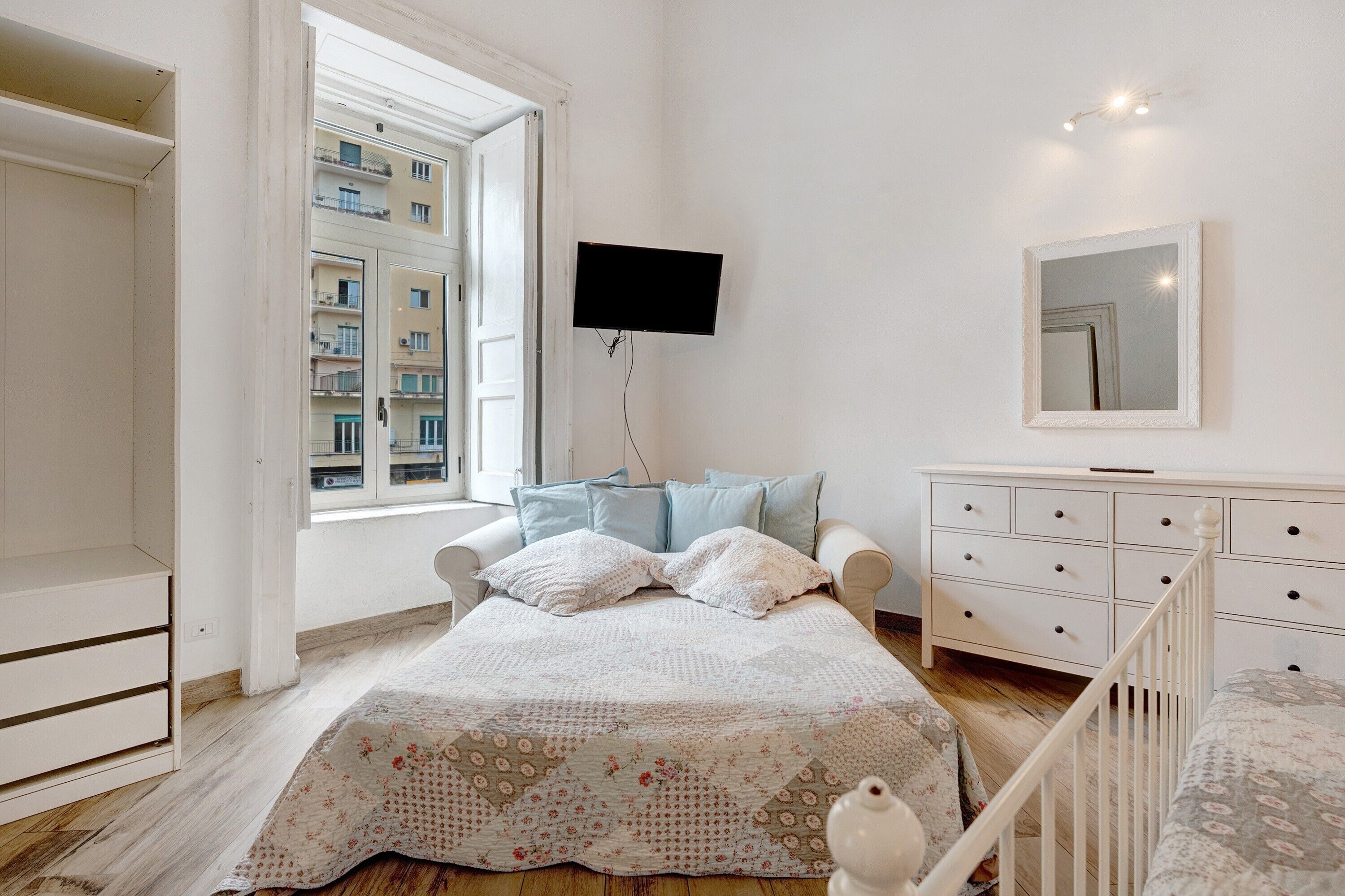 A bedroom with a bed, a dresser, a TV, and a window with a view of buildings.