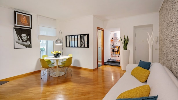 A modern living room with a white sofa, a dining table with chairs, and a kitchen visible through an open door.