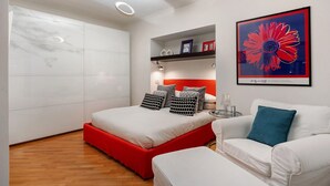 A modern bedroom with a bed, a sofa, a red accent wall, and a framed artwork.
