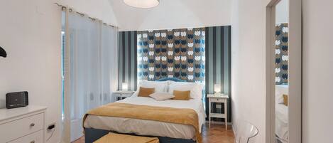 A bedroom with a bed, bedside tables, a desk, a chair, a hanging light fixture, and a mirror.