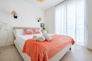 A neatly made bed with a red blanket, two pillows with a cartoon face design, and two rolled towels. There are two wall-mounted lamps, a small plant on a nightstand, and a window with sheer curtains.
