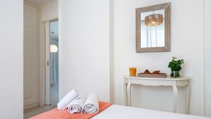 Hotellihuone, jossa on sänky, valkoinen konsolipöytä peilillä, maljakko, jossa on kasvi, ja ovi, joka johtaa toiseen huoneeseen.