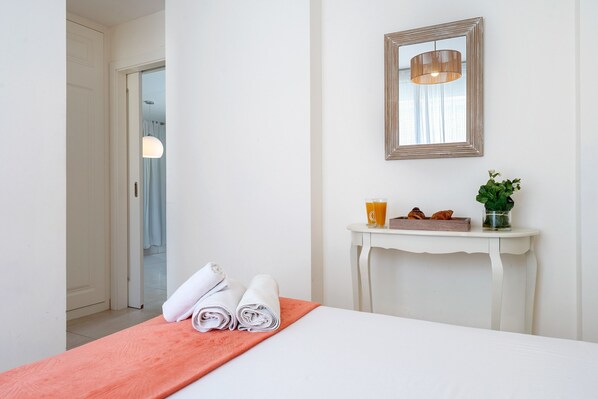 Una habitación de hotel con una cama, una consola blanca con espejo, un jarrón con una planta y una puerta que da a otra habitación.