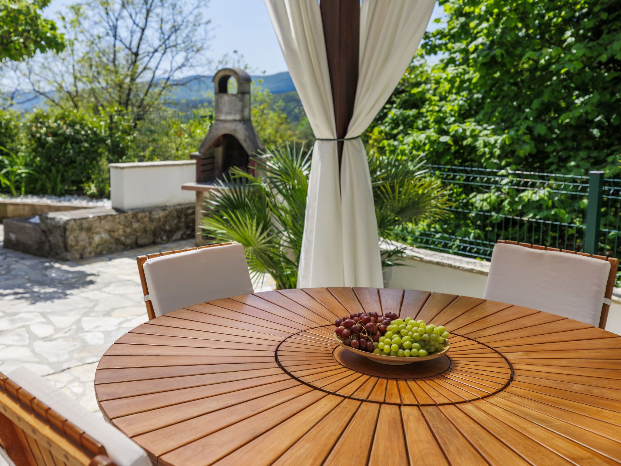 A round wooden table with a bowl of grapes, surrounded by chairs, set outdoors with a view of greenery and a stone wall in the background.