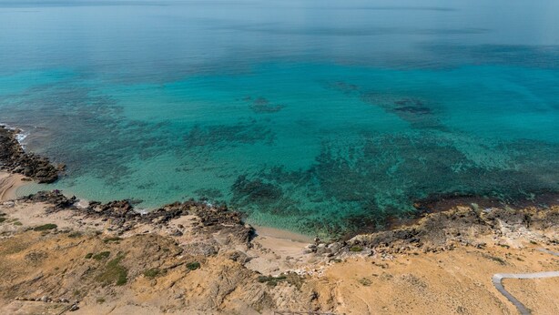 Vista aérea de una zona costera con aguas turquesas cristalinas, terreno rocoso y un camino visible que lleva a una playa.