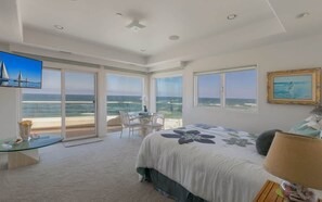 Une chambre avec un lit, un bureau et une chaise, de grandes fenêtres donnant sur la mer, et un téléviseur fixé au mur.