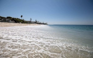 Una playa con agua cristalina, orilla arenosa y una vista distante de una zona residencial con árboles y edificios.