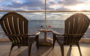 Dos sillas de madera y una mesa pequeña con dos copas de vino tinto en un balcón con vistas al mar.