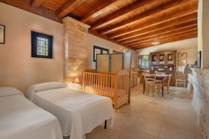 A room with two beds, a crib, a dining table with chairs, a cabinet with glass doors, and a stone wall.