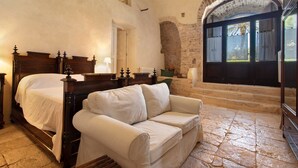 A bedroom with a double bed, a white sofa, stone flooring, a stone wall, and a view through a window.