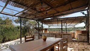 An outdoor patio with a wooden table and chairs, a thatched roof, stone flooring, and a view of trees and hills.