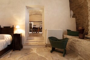 Une chambre à coucher comprenant un lit, une table de chevet, une lampe, une chaise en osier, un radiateur et un mur en pierre.