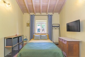 A bedroom with a bed, a TV, a wooden cabinet, a metal shelf unit, and a window with curtains.