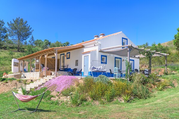 A white building with blue accents, a covered outdoor seating area, and a garden with various plants.