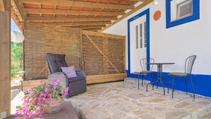 Een overdekt terras met stenen vloer, een rieten stoel, een comfortabele bank en een tafel met stoelen.
