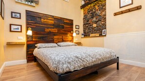 Une chambre avec une tête de lit en bois, un lit recouvert d’une literie à motifs, un mur en pierre et une étagère ornée de photos encadrées.