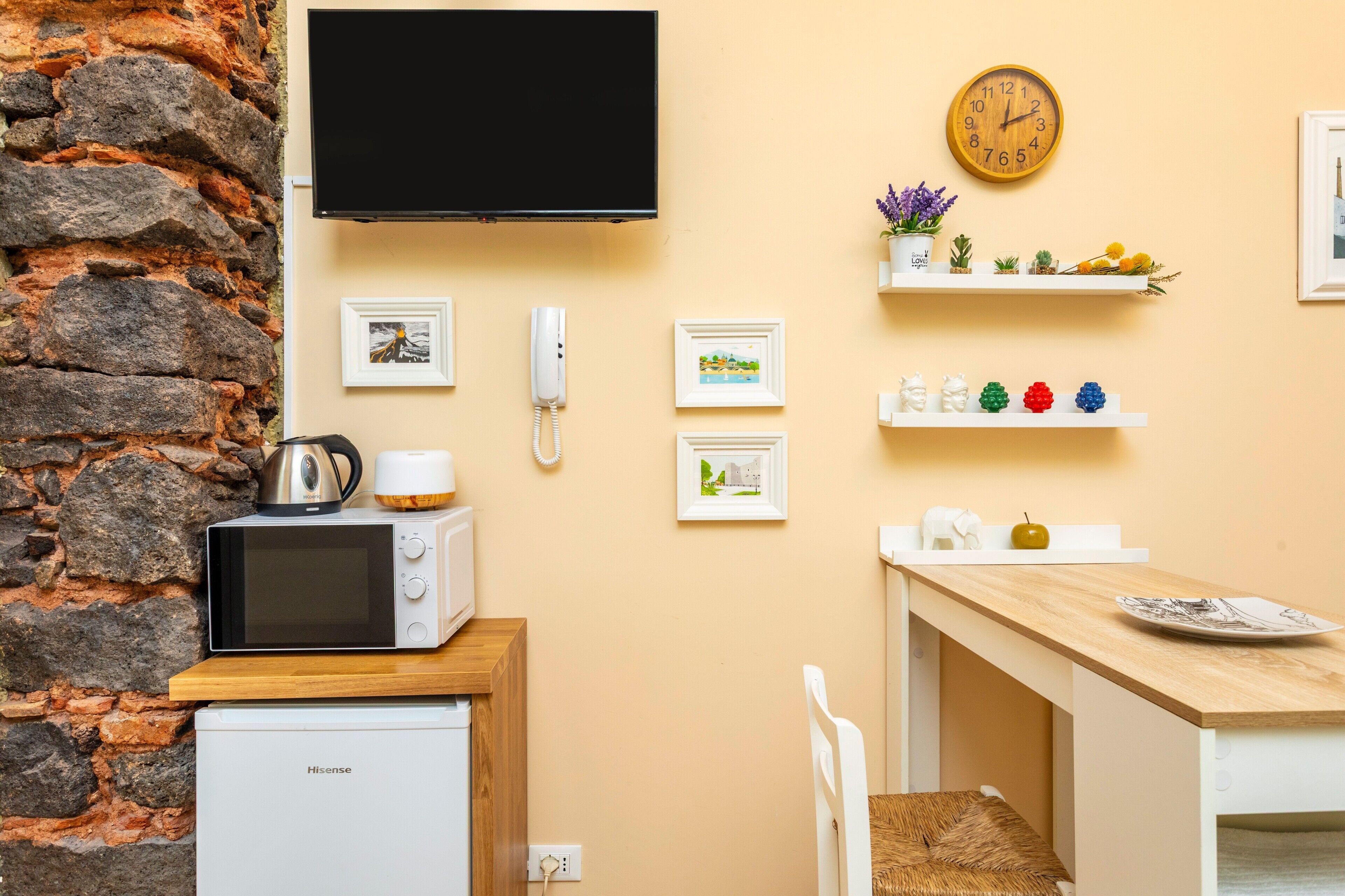 Eine kompakte Küche mit Mikrowelle, kleinem Kühlschrank, Holzausstattung, einem an der Wand montierten Fernseher und einer Uhr.