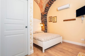 Un dormitorio con una cama blanca, un arco de piedra, un equipo de aire acondicionado, un televisor montado y un estante de madera.