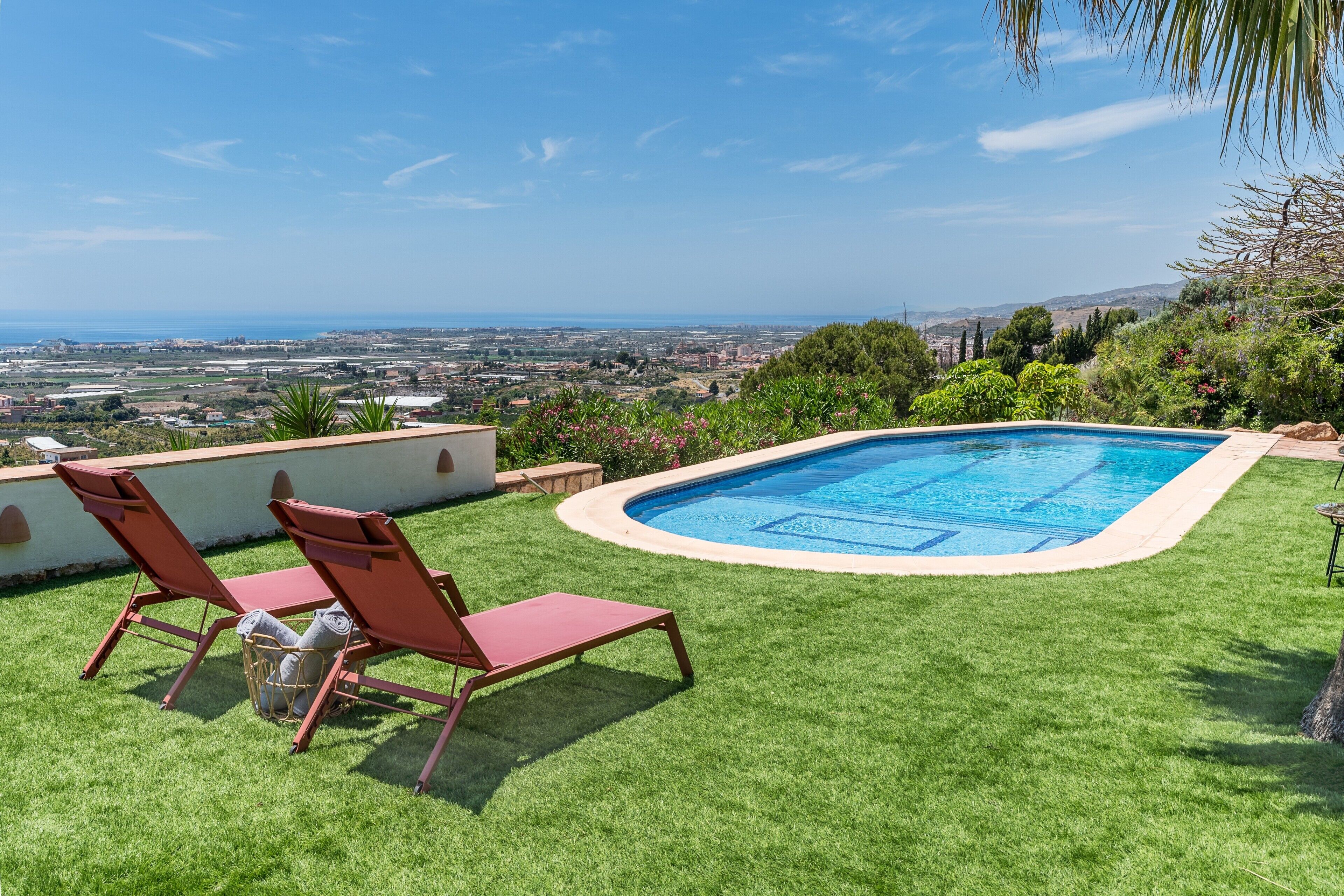 Una piscina con dos sillas reclinables rojas, un césped y vistas a un paisaje urbano.