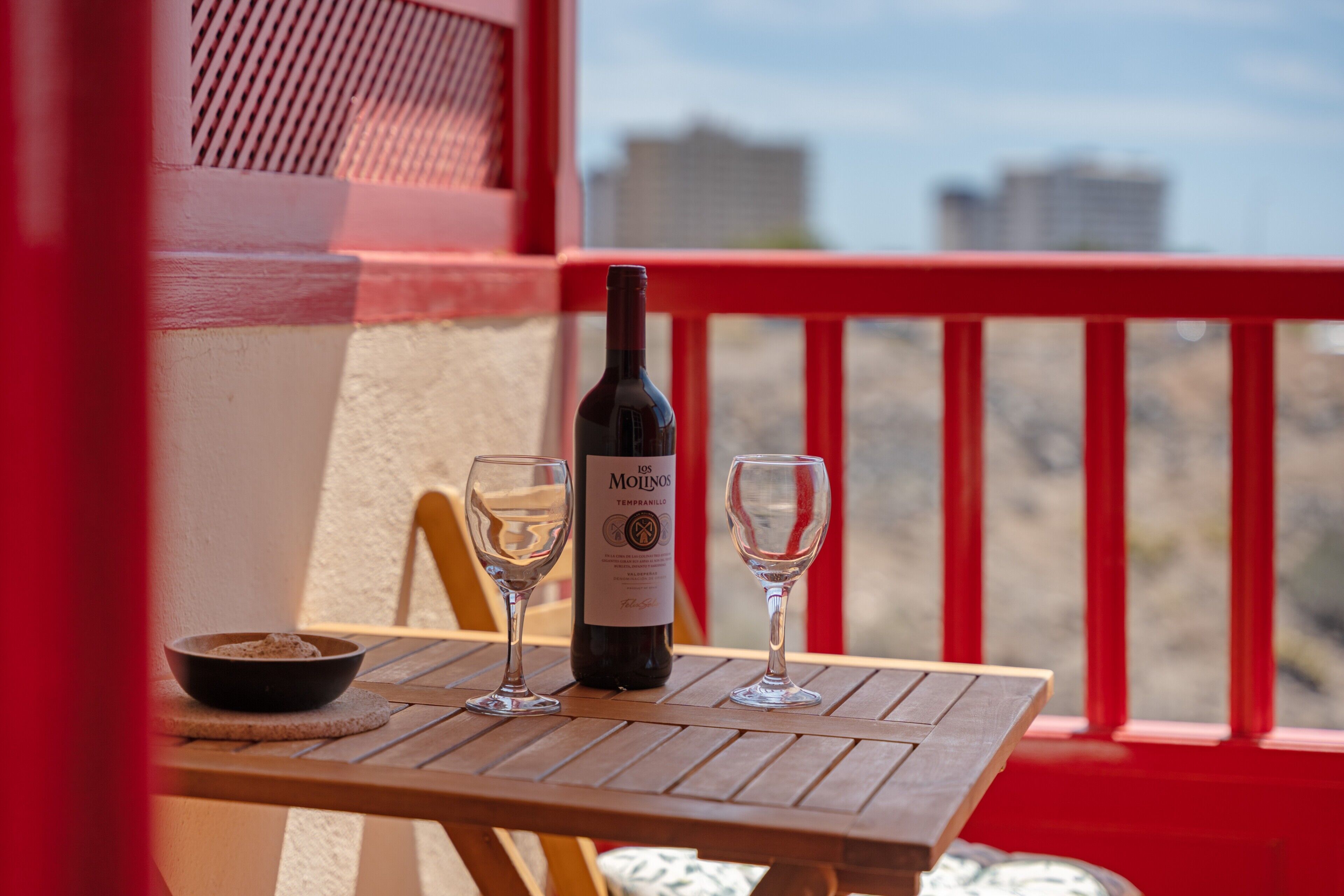 A table with a bottle and two glasses on a balcony with a red railing and a view of buildings.