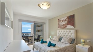 A bedroom with a bed, a nightstand, a lamp, a painting, a desk, and a chair.
