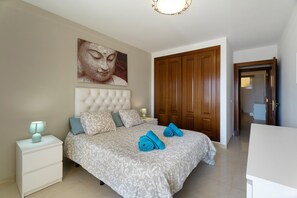 Un dormitorio con cama, mesita de noche, decoración en la pared, armario de madera y una puerta que da a otra habitación.