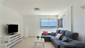 A modern living room with a flat-screen TV, a grey sofa, a white coffee table, and a view of the sea through a window.