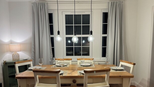 Comedor con mesa y sillas de madera, una lámpara y un ventanal con cortinas.