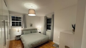 Un dormitorio con cama, lámparas de noche, una ventana con cortinas, un radiador y suelo de madera.