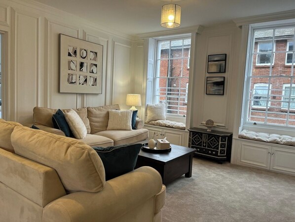 A living room with a beige sofa, a coffee table, and a lamp. There are windows with a view of a brick building. - Spacious 2B Flat in Clement House (Hertfordshire)