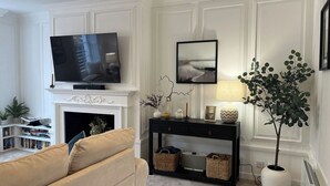Uma sala de estar com lareira, televisão, sofá, mesa de console com objetos decorativos e um vaso de planta.