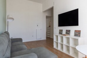 A modern living room with a grey sofa, a white lamp, a flat-screen TV, and a shelf with signs indicating famous locations.