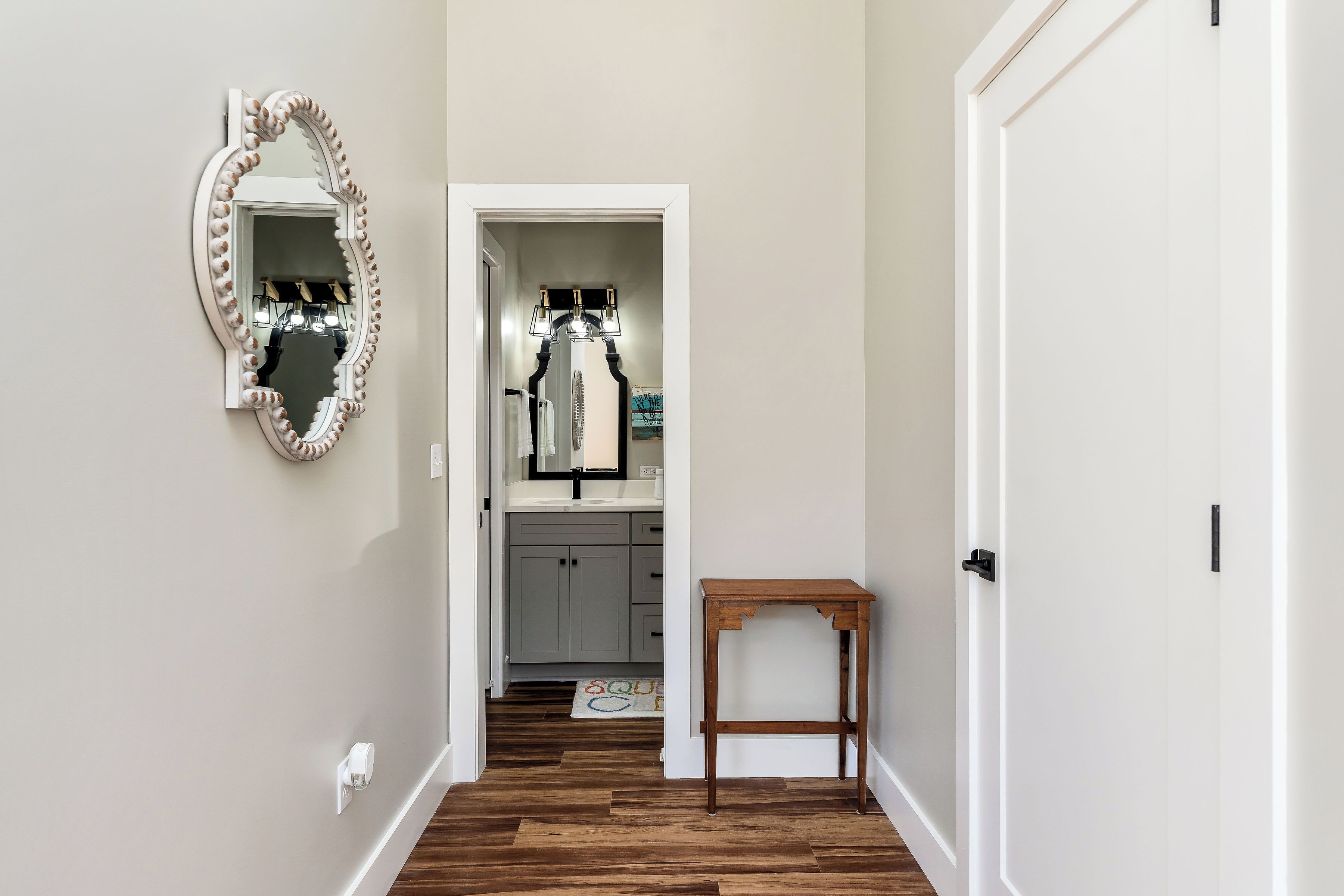 A hallway with a wooden floor, a mirror with a decorative frame, a small wooden table, and a door.
