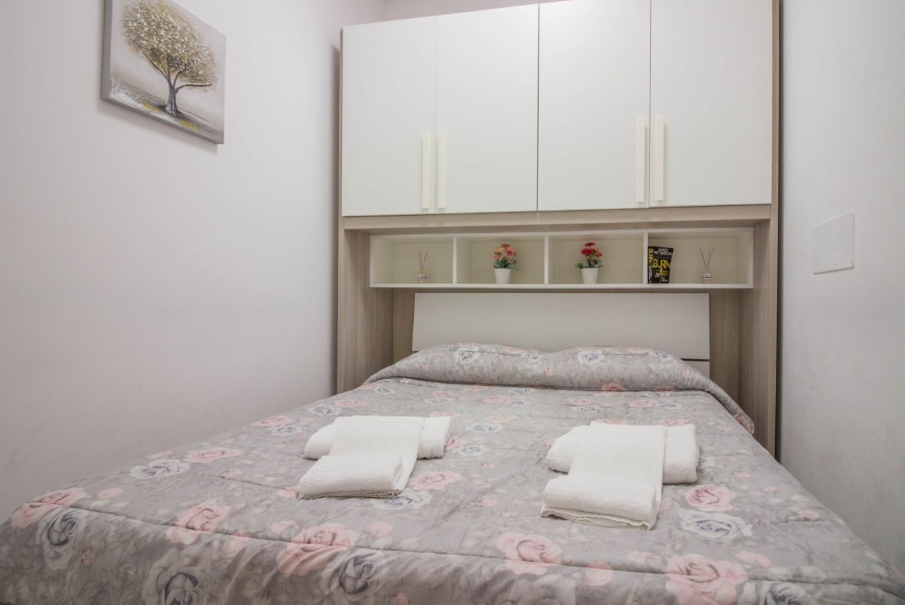 A bedroom with a bed, a nightstand, a wardrobe, and a wall-mounted shelf with decorative items.