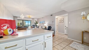 A kitchen with white cabinets and gold handles, a countertop with a red towel, and a view of the living area.