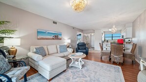 A spacious living room with a white sofa, a dining area with wicker chairs, and a kitchen in the background.