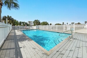 Ein Swimmingpool mit Holzterrasse, weißem Zaun und Palmen im Hintergrund.