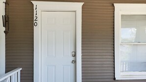 Une porte blanche avec le numéro 1120 inscrit sur le mur à côté, une fenêtre avec des stores et un petit espace de véranda.
