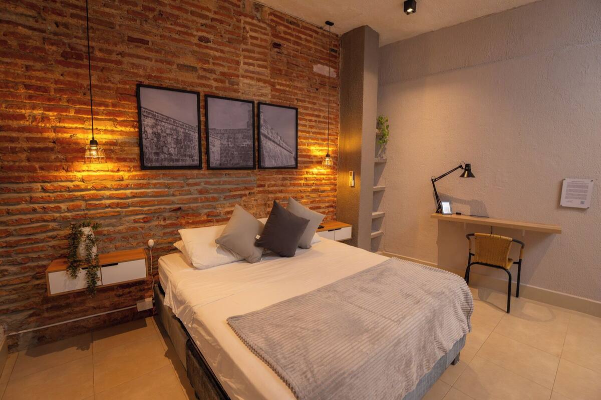 A modern bedroom with a brick wall, a bed with pillows, a desk, and a chair.