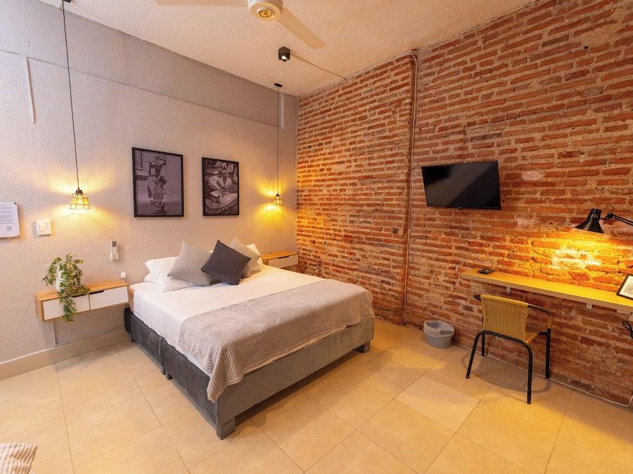 Un dormitorio moderno con pared de ladrillo, una cama con cabecera gris, un escritorio con silla y un televisor de pantalla plana montado en la pared.