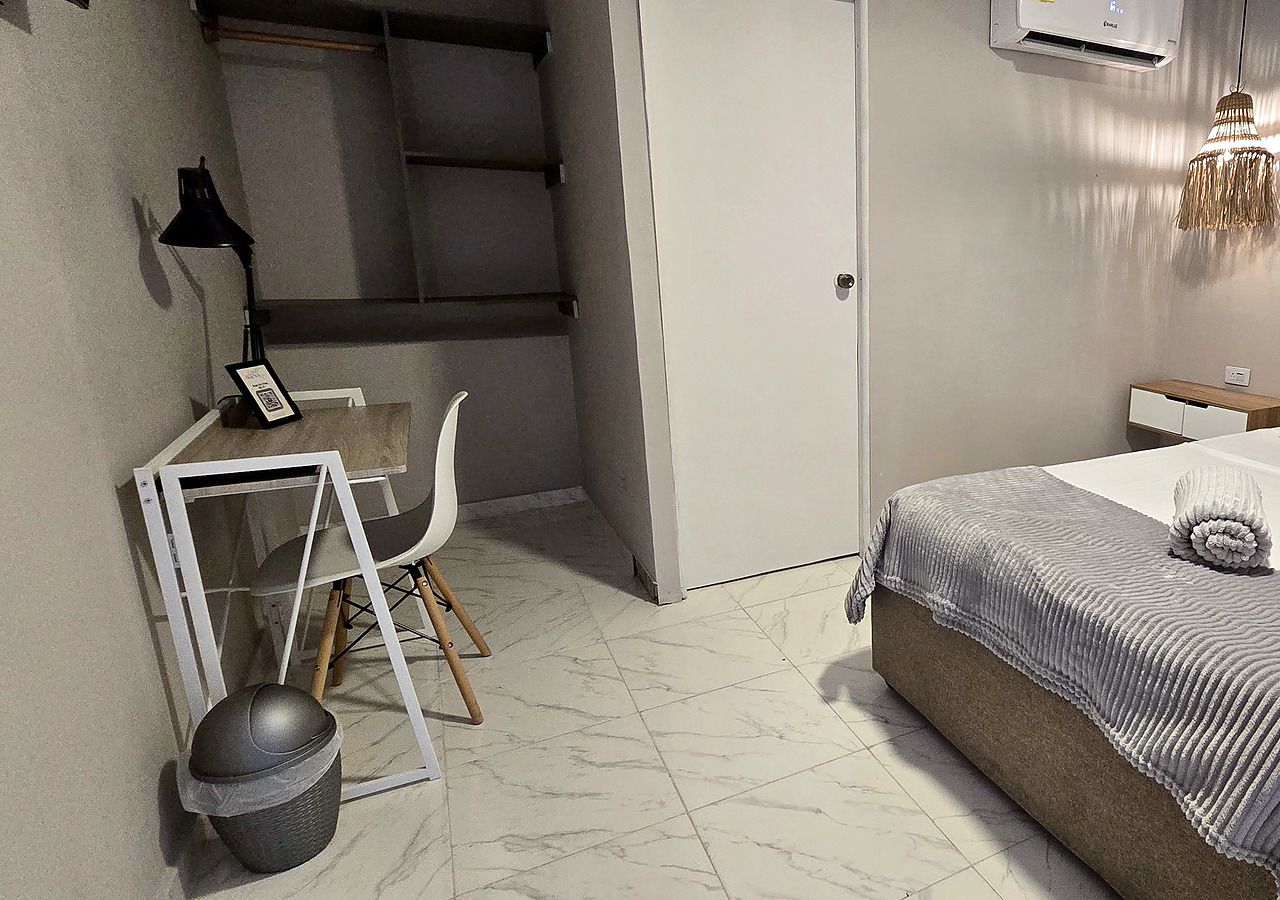 Ein kleines Schlafzimmer mit einem Bett, einem Schreibtisch mit Stuhl, einer kleinen Mülltonne und einer Wand-Klimaanlage.