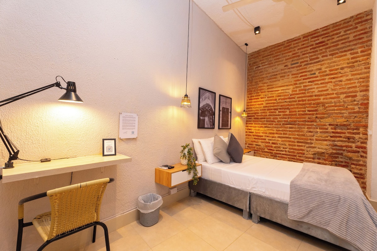 A bedroom with a brick wall, a bed, a desk with a lamp, a chair, and a small trash bin.